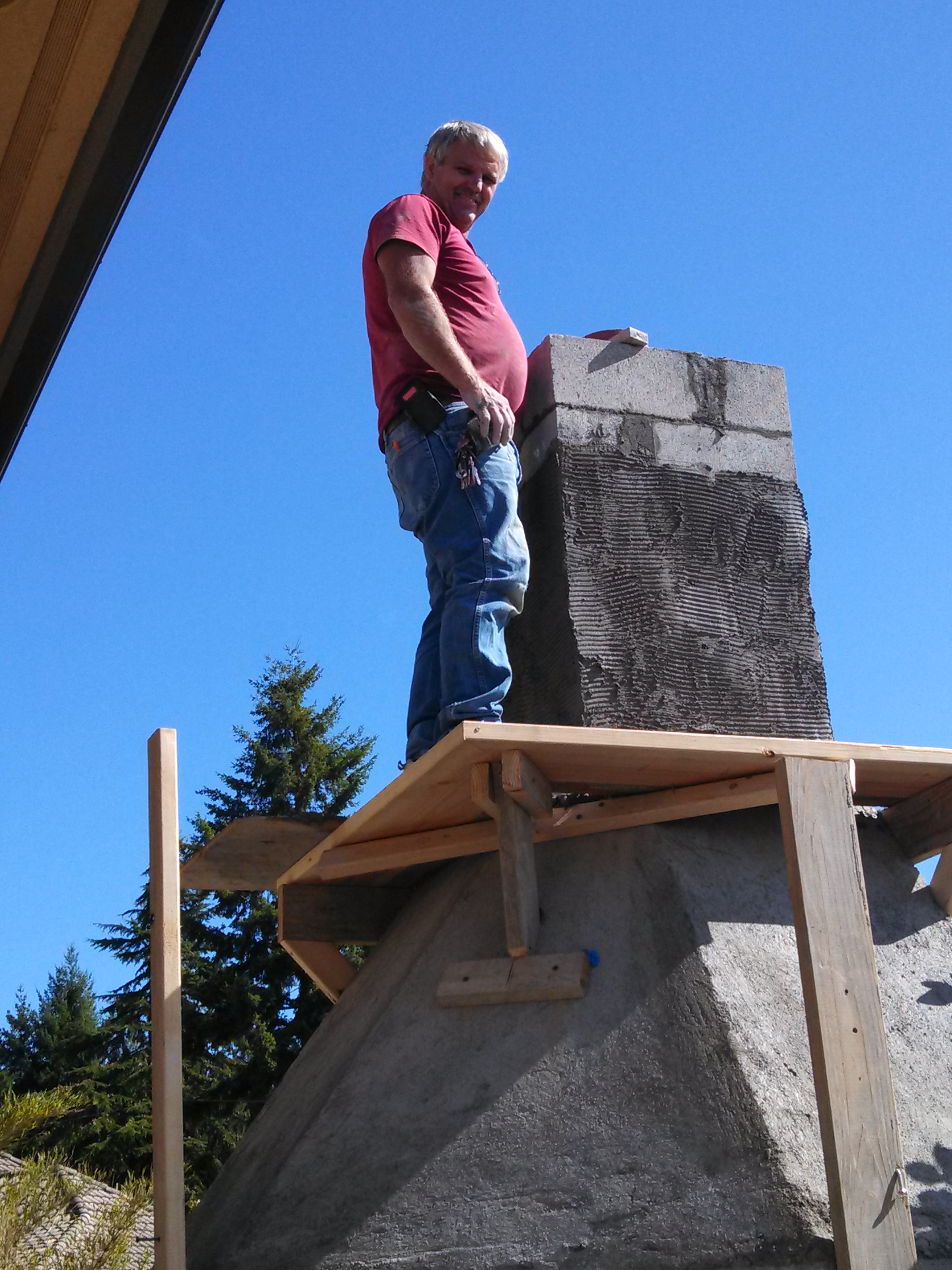A man standing on a wooden platform working with a large stone structure, with a clear blue sky and trees in the background.